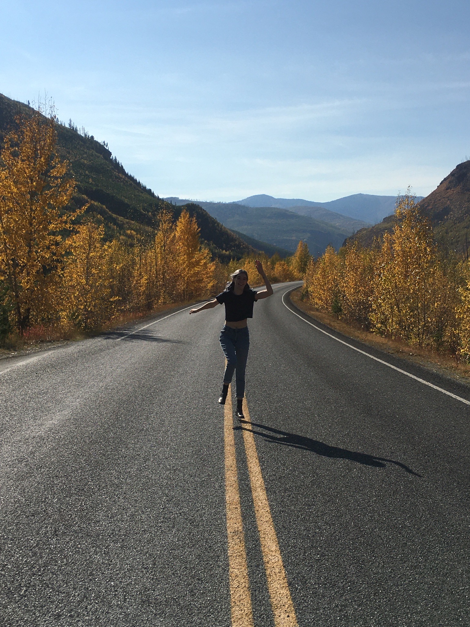 Julia skipping down a road in Montana. The trees lining the road are a bright yellow and there are hills in the background.